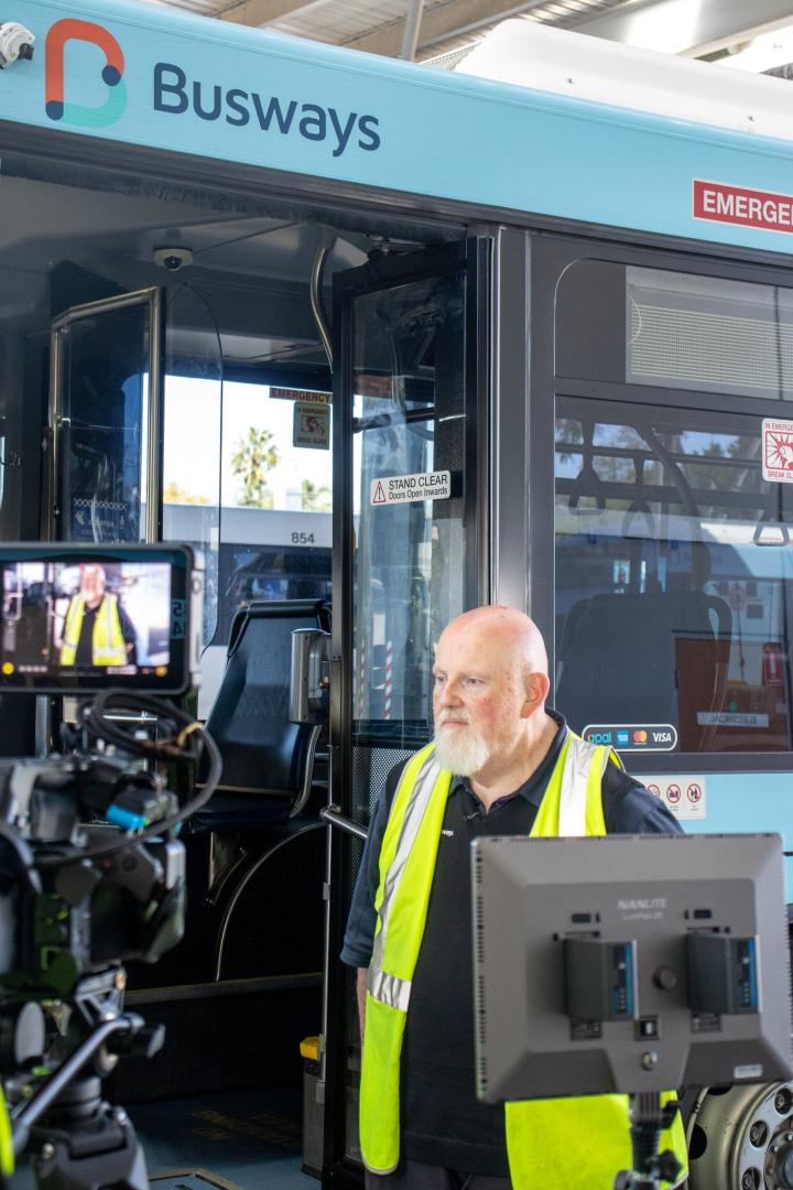 Bus driver standing in front of a blue bus being filmed